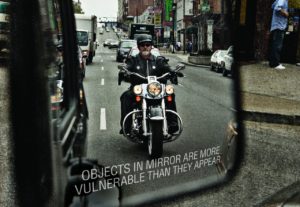 Motorcyclist approaching in vehicle mirror. Text on bottom of mirror reads, Objects in mirror are more vulnerable than they appear.